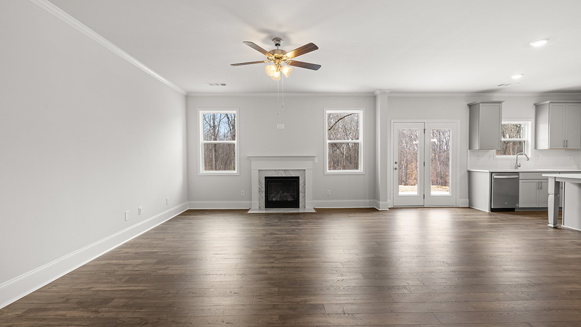 Spacious modern living room with hardwood floors, light gray walls, and white trim. Includes a ceiling fan, fireplace, and two large windows. Kitchen with gray cabinets and stainless steel dishwasher visible on the right.