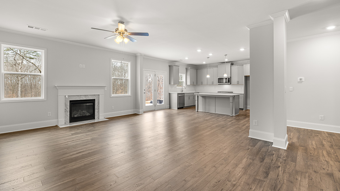 Spacious open-concept living area with hardwood floors, light gray walls, and ceiling fan. Features a modern kitchen with island and stainless steel appliances, adjacent to a cozy fireplace.