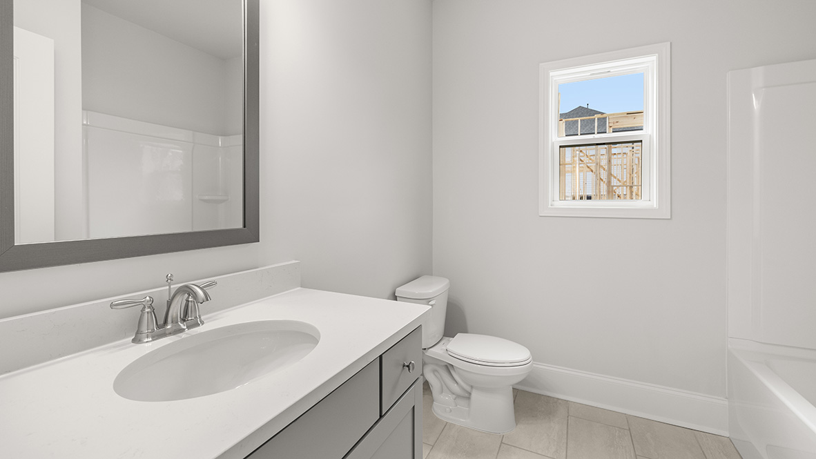 Modern bathroom with white walls and large gray framed mirror above a white sink. A small window shows a partial view of a building under construction.