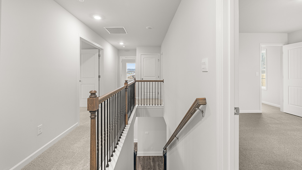 Hallway with beige carpet, white walls, and wooden railing with black spindles. Open doors lead to rooms, and natural light filters from windows. Modern and calm.