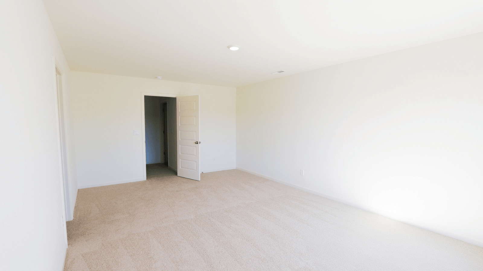 Empty room with beige carpet and white walls, featuring an open door leading to a darker hallway.