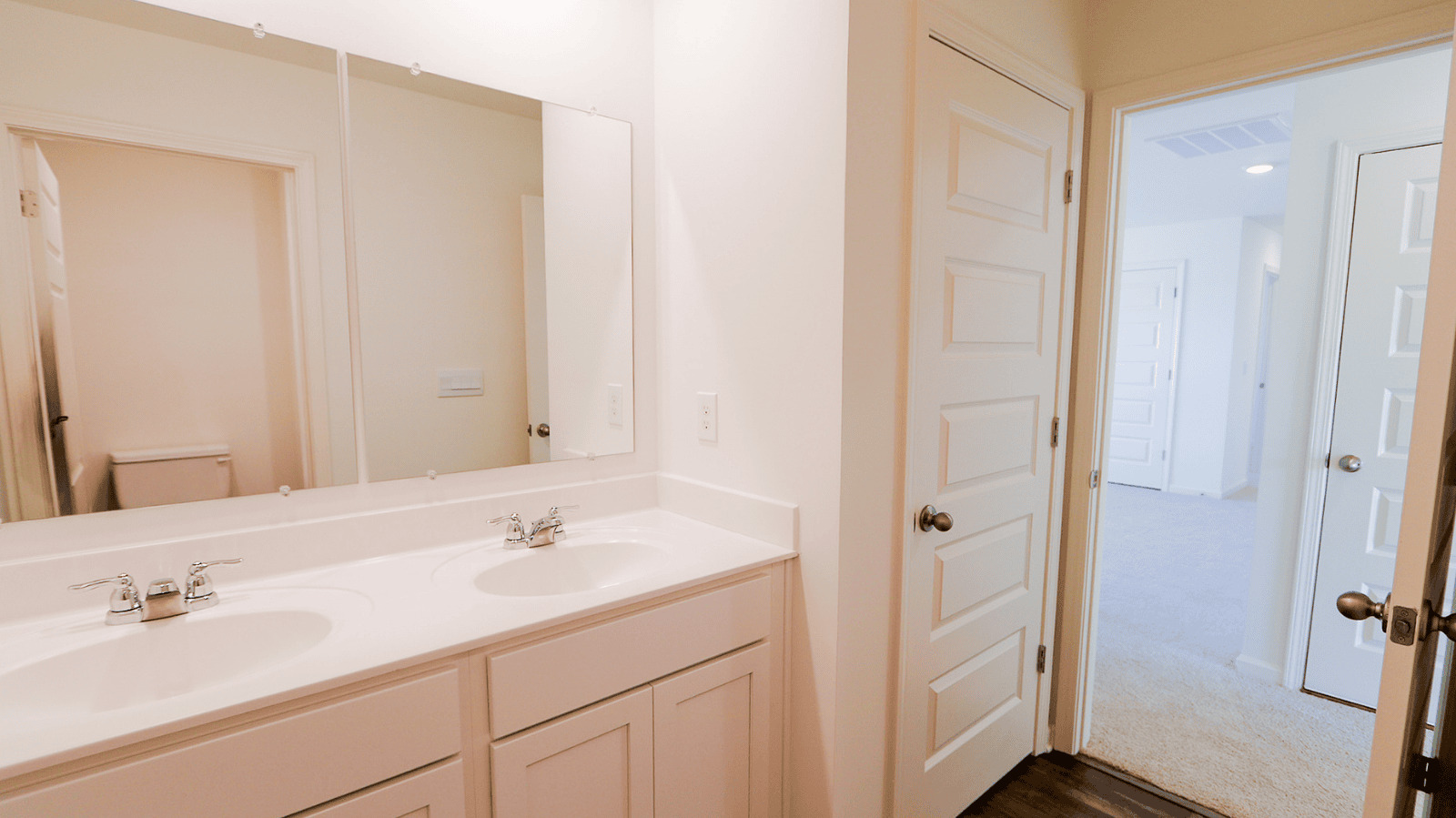 Modern double vanity with sink and mirrors in a well-lit bathroom, leading to a hallway and additional doors.