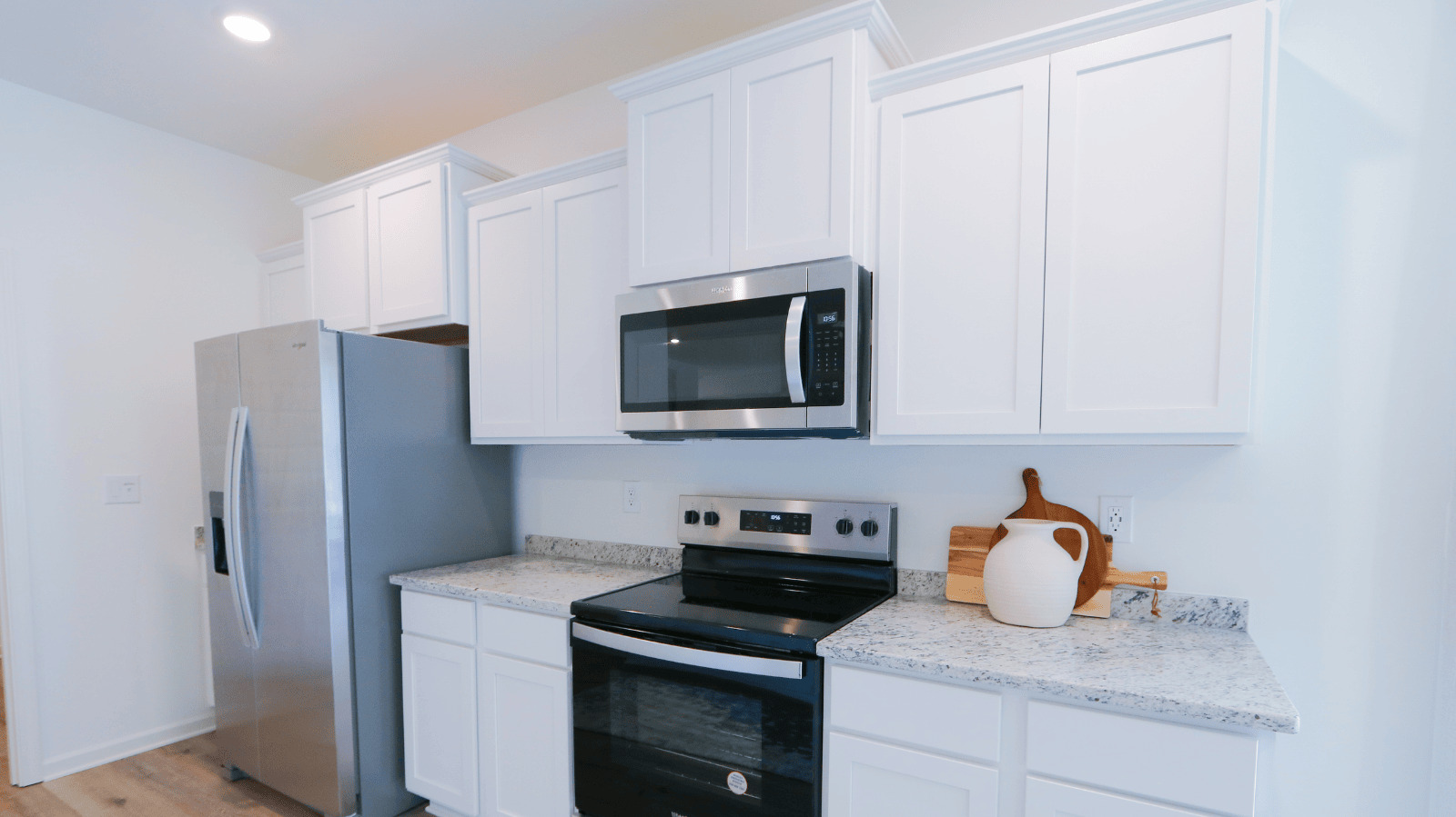 Modern kitchen with white cabinets and stainless steel appliances, featuring a side-by-side refrigerator and built-in microwave. The granite countertop is decorated with a white vase of wheat, a teapot, cups, and a bowl of nuts, creating a stylish and inviting cooking space.