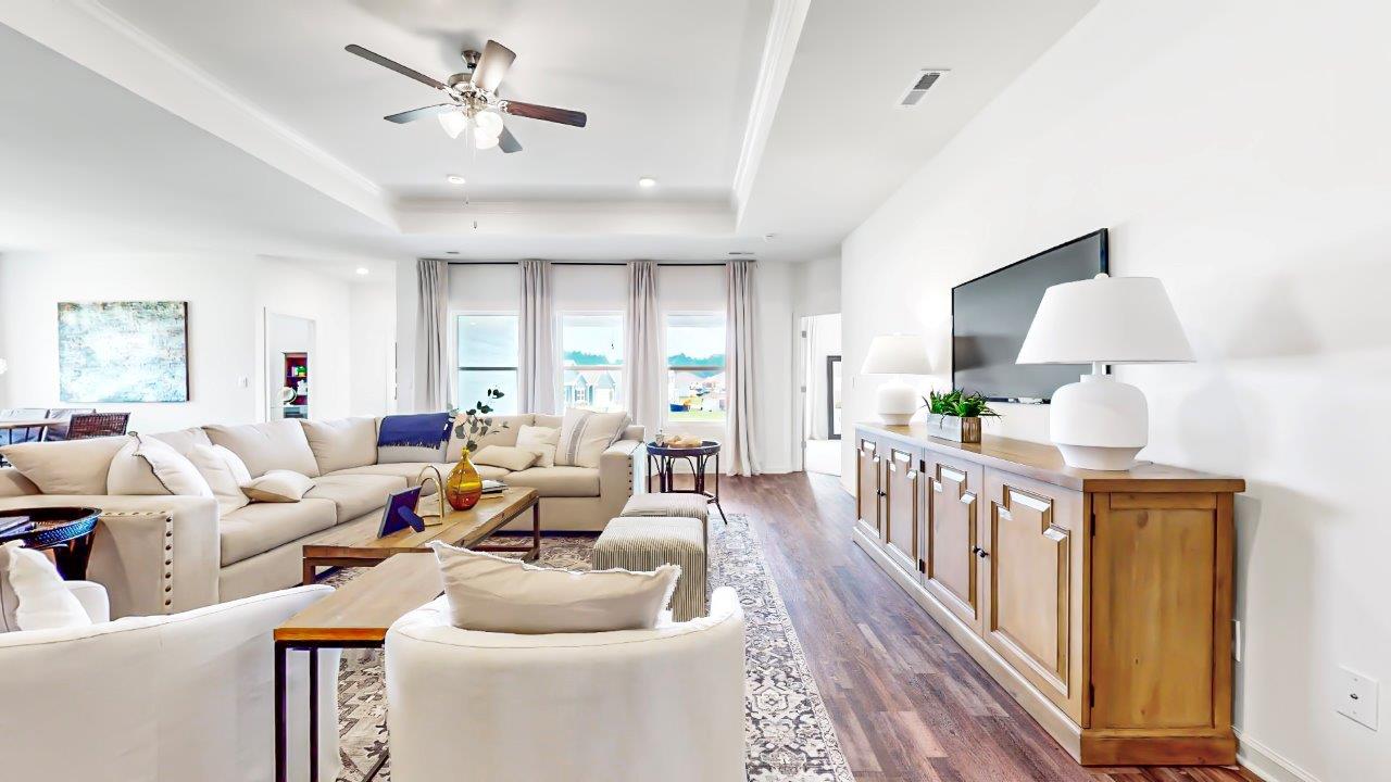 Open concept living room shows the trey ceilings as well as a large sectional couch, coffee table, media consol with tv and 2 accent chairs