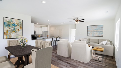 n open living area with light wood flooring, neutral-colored walls, and a ceiling fan with lights in the center of the room. The space is bright and airy, with natural light streaming in from large windows on one side. Multiple doors lead to other rooms, contributing to the open-concept layout that connects the living area seamlessly with the kitchen.