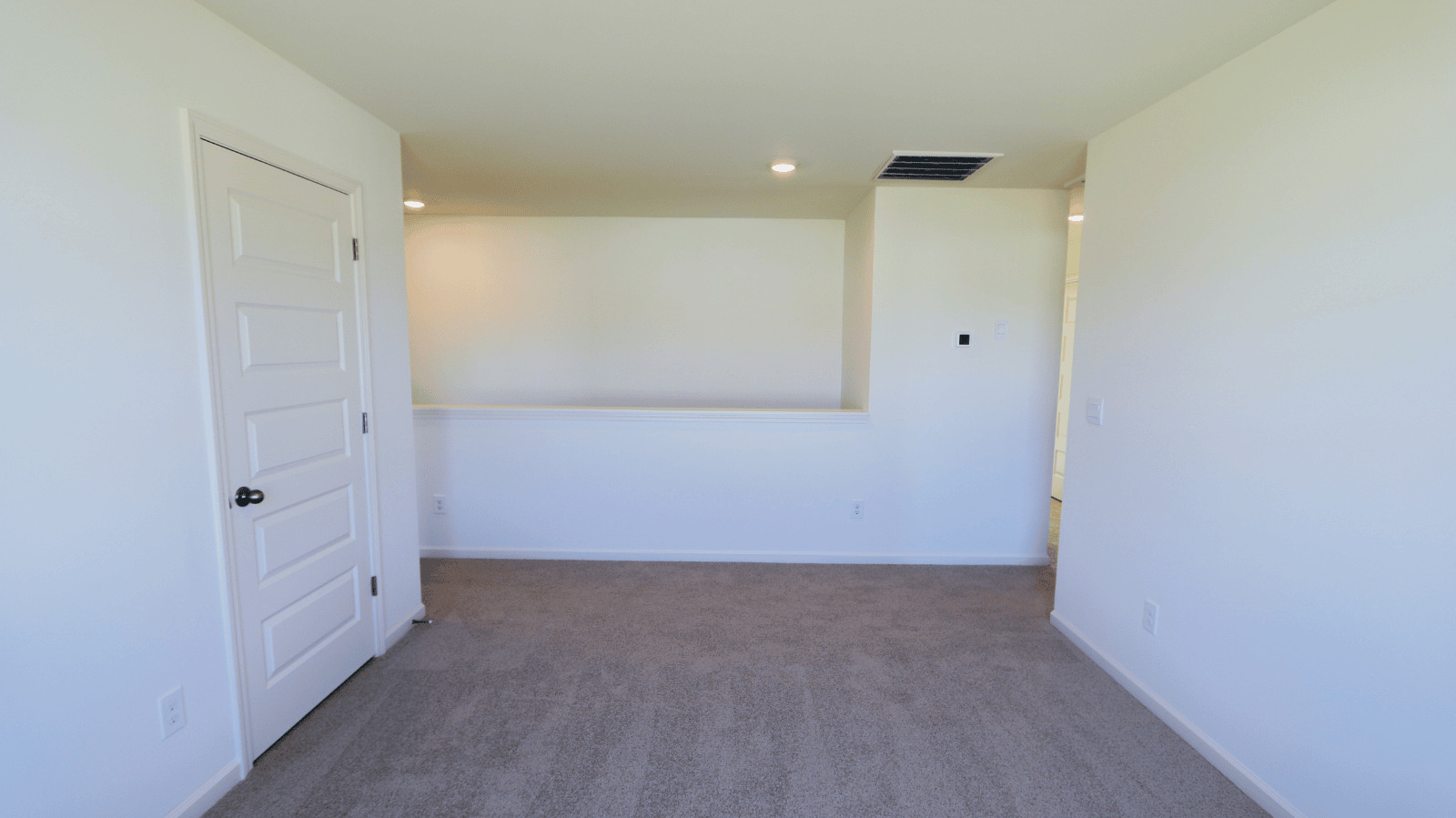 Empty room with light-colored walls, carpet flooring, a closed door on the left, and a small opening to another space.