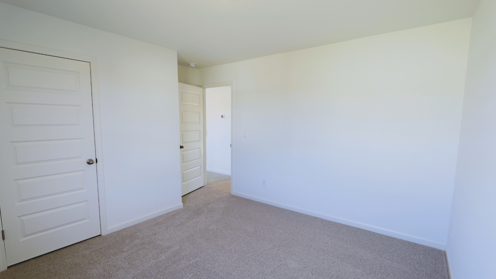 Empty room with white walls and carpet, featuring two doors and natural light. Ideal for various layouts.