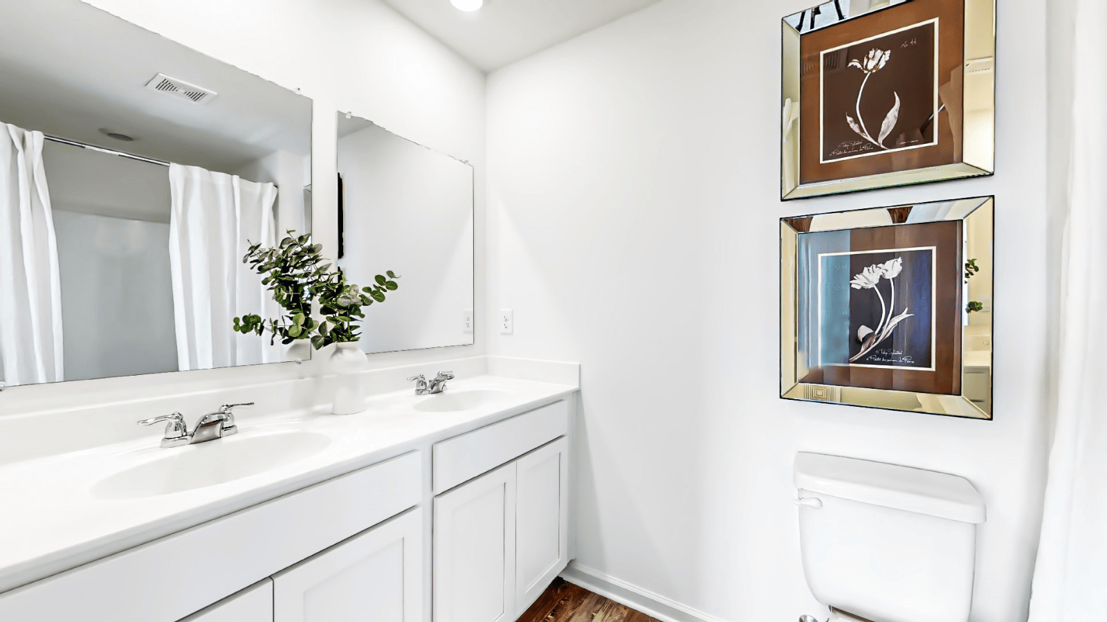A bathroom view from the door featuring white cabinets, a double vanity, a shower on the right, and a toilet in the center.