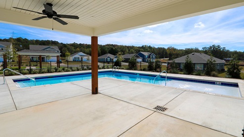 View of a swimming pool surrounded by a patio and residential buildings, with a covered area and clear blue sky above.
