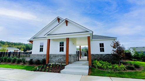 A modern, single-story house with white siding and wooden accents, surrounded by greenery and a clear blue sky.