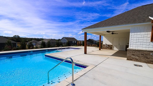 A clear blue swimming pool beside a covered patio. The sky is partly cloudy, with houses visible in the background.
