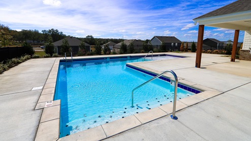 A clear blue pool surrounded by a concrete deck, with a gentle slope and steps, set against a backdrop of houses and trees.