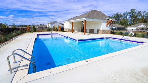 A clear blue swimming pool with steps, surrounded by a concrete deck and a small pavilion in the background under a blue sky.