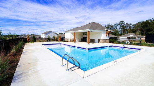 A serene outdoor pool area featuring a clear blue pool, a covered seating area, and surrounding greenery under a bright sky.