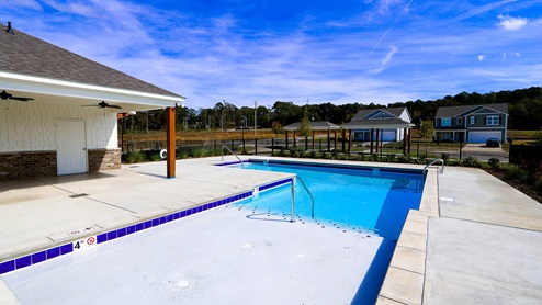 A sunny pool area featuring a clear blue pool beside a building, with homes visible in the background and clear skies above.