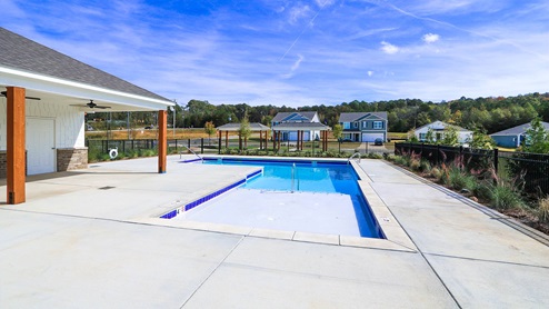 A spacious pool area featuring clear water, surrounded by concrete and a wooden fenced patio against a blue sky.