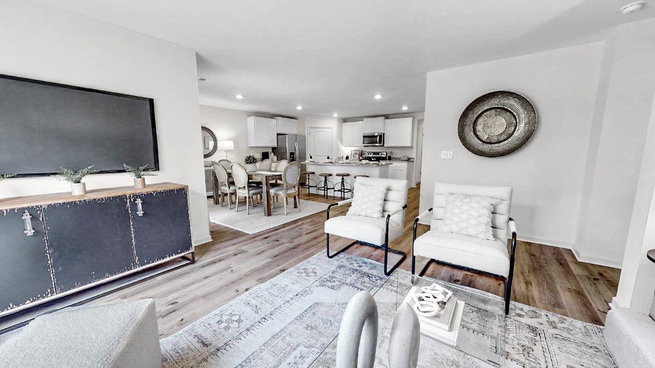 A modern open-concept living room and kitchen with a neutral color palette. The space features a comfortable seating area, round dining table, and stylish decor including a large wall-mounted TV and a decorative wall piece. The flooring is a light wood finish.