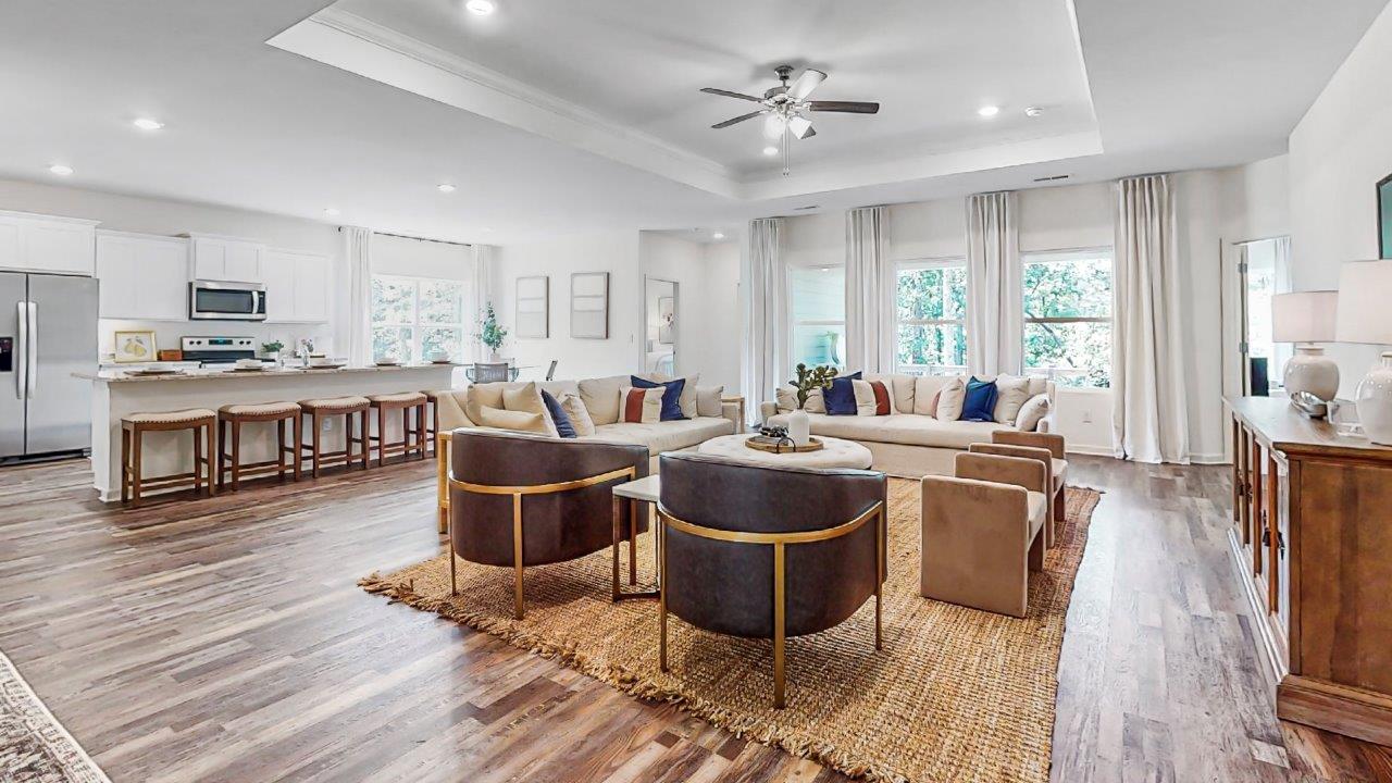 Modern open-concept living room with hardwood floors, ceiling fan, and large windows. Features a beige sofa, armchairs, and a kitchen with a bar counter and stools in the background.