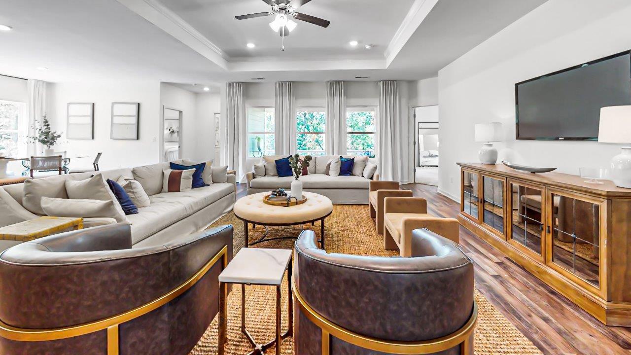 Spacious living room with two large sofas, leather chairs, a wooden coffee table, a TV on a console, and large windows. The room is decorated in neutral tones with blue accents.