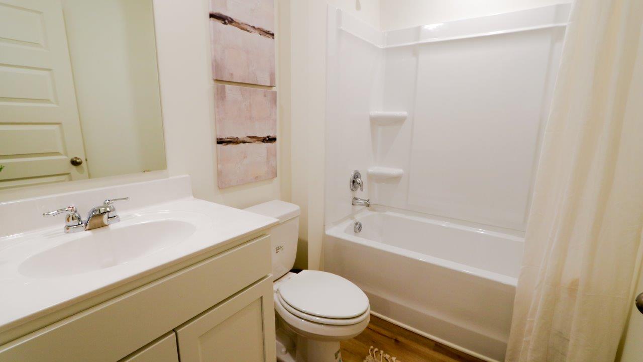The bathroom is comprised of a tub/shower, toilet, and vanity. The walls are white, and the floors are made of wood, which gives the space a clean and natural aesthetic.