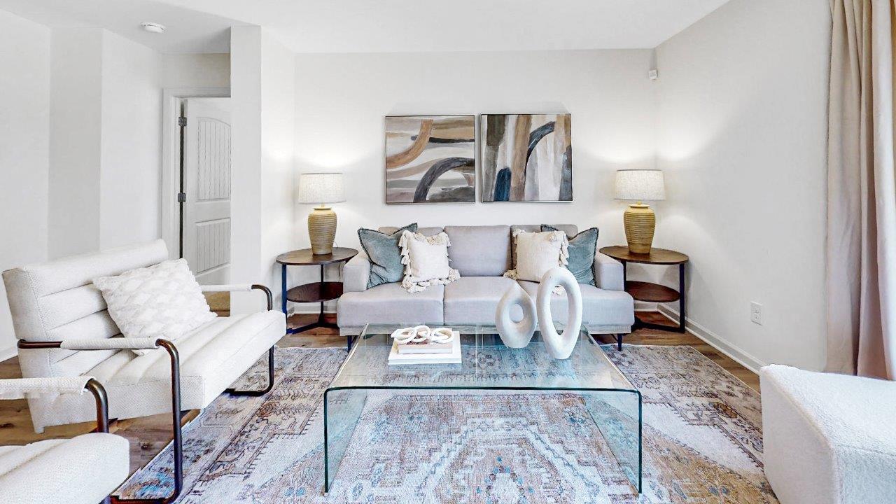A modern living room with a light gray sofa adorned with pillows, accompanied by two armchairs. A glass coffee table sits atop a patterned rug. Two abstract paintings hang above the sofa, flanked by lamps on round wooden side tables.