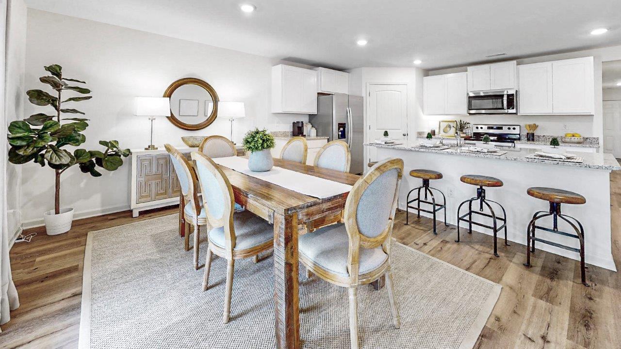 A modern dining room and kitchen with a wooden table and six chairs on a light rug. The kitchen features white cabinets, stainless steel appliances, and a breakfast bar with three stools. A round mirror hangs above a decorative cabinet.