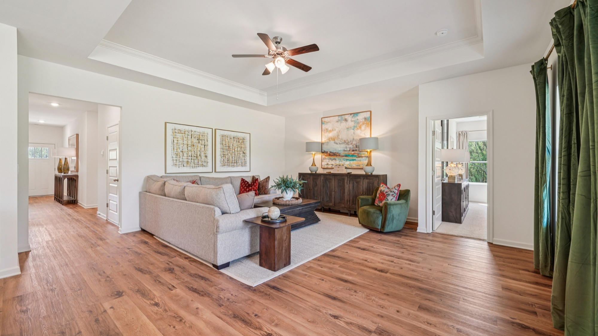 A modern living room with light wood floors and white walls. It features a sofa, armchair, and two ottomans, facing a large TV on a wooden console. Large windows and a glass door provide natural light. Neutral decor complements the space.