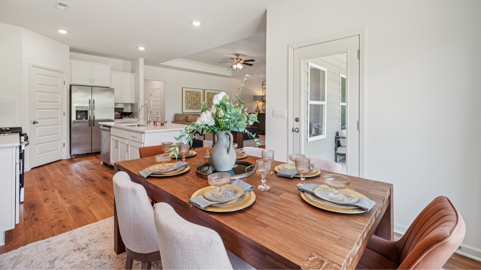 A modern dining room and kitchen with a wooden table and six chairs on a light rug. The kitchen features white cabinets, stainless steel appliances, and a breakfast bar with three stools. A round mirror hangs above a decorative cabinet.