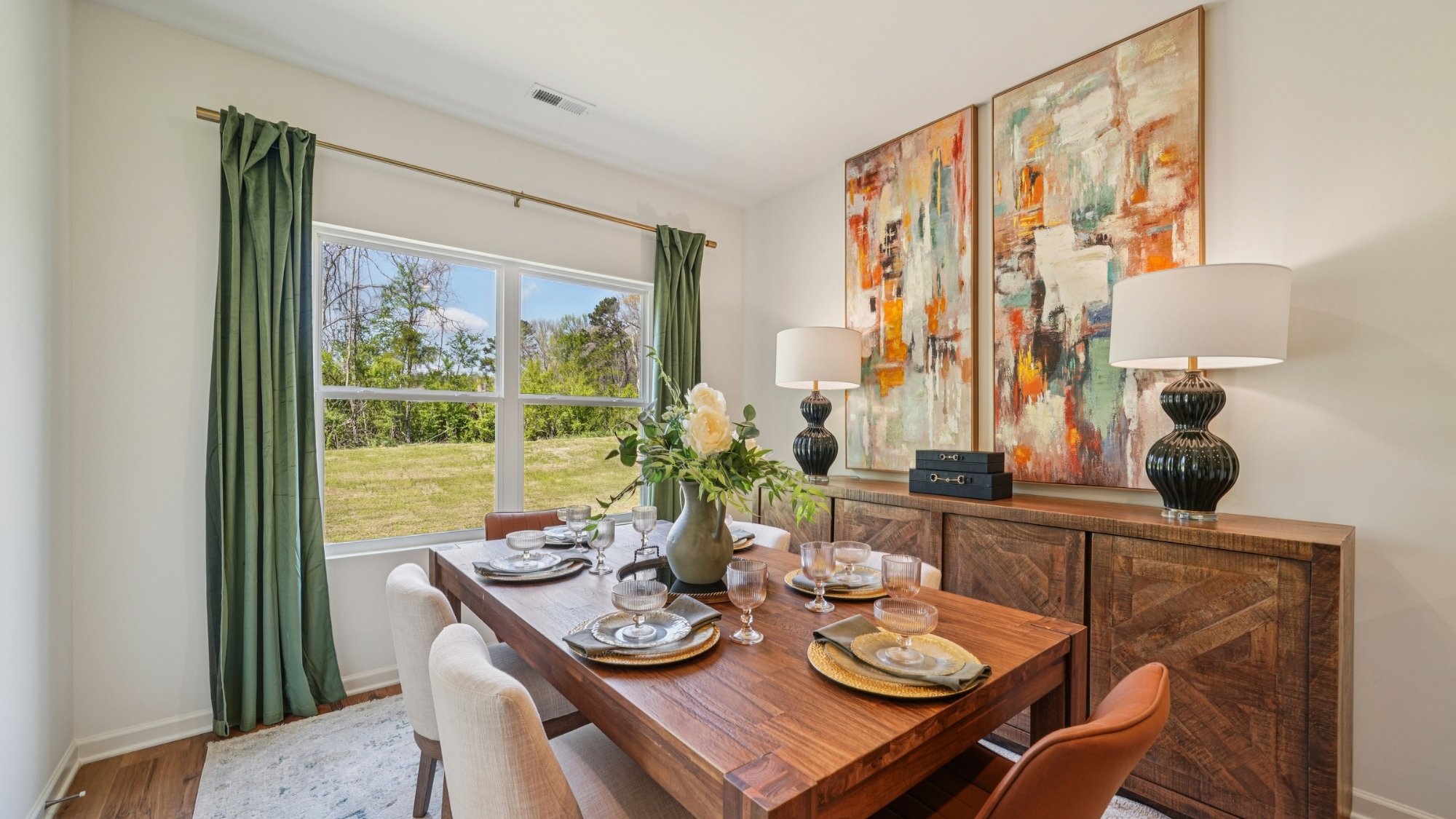 A modern dining room and kitchen with a wooden table and six chairs on a light rug. The kitchen features white cabinets, stainless steel appliances, and a breakfast bar with three stools. A round mirror hangs above a decorative cabinet.