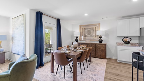 A modern dining room and kitchen with a wooden table and six chairs on a light rug. The kitchen features white cabinets, stainless steel appliances, and a breakfast bar with three stools. A round mirror hangs above a decorative cabinet.