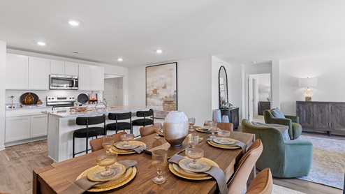 A modernA modern dining room and kitchen with a wooden table and six chairs on a light rug. The kitchen features white cabinets, stainless steel appliances, and a breakfast bar with three stools. A round mirror hangs above a decorative cabinet. living room with light wood floors and white walls. It features a sofa, armchair, and two ottomans, facing a large TV on a wooden console. Large windows and a glass door provide natural light. Neutral decor complements the space.