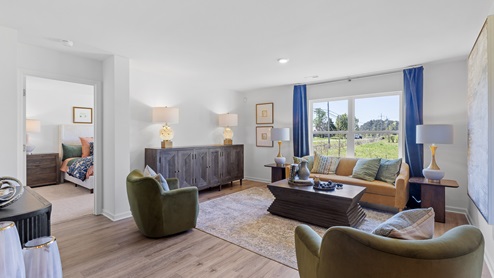 A modern living room with light wood floors and white walls. It features a sofa, armchair, and two ottomans, facing a large TV on a wooden console. Large windows and a glass door provide natural light. Neutral decor complements the space.