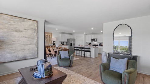 A modern living room with light wood floors and white walls. It features a sofa, armchair, and two ottomans, facing a large TV on a wooden console. Large windows and a glass door provide natural light. Neutral decor complements the space.