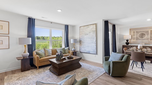 A modern living room with light wood floors and white walls. It features a sofa, armchair, and two ottomans, facing a large TV on a wooden console. Large windows and a glass door provide natural light. Neutral decor complements the space.