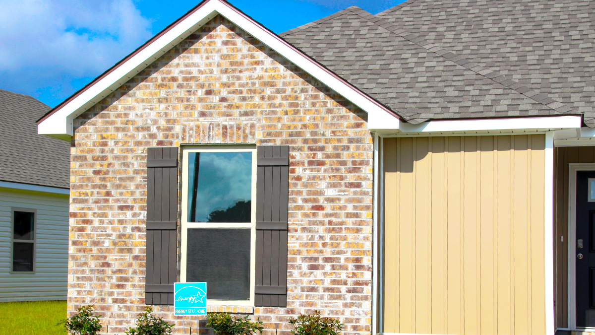 Single-story home with brown brick and tan vinyl hardie. Black glass front door with brown shutters and white trim.