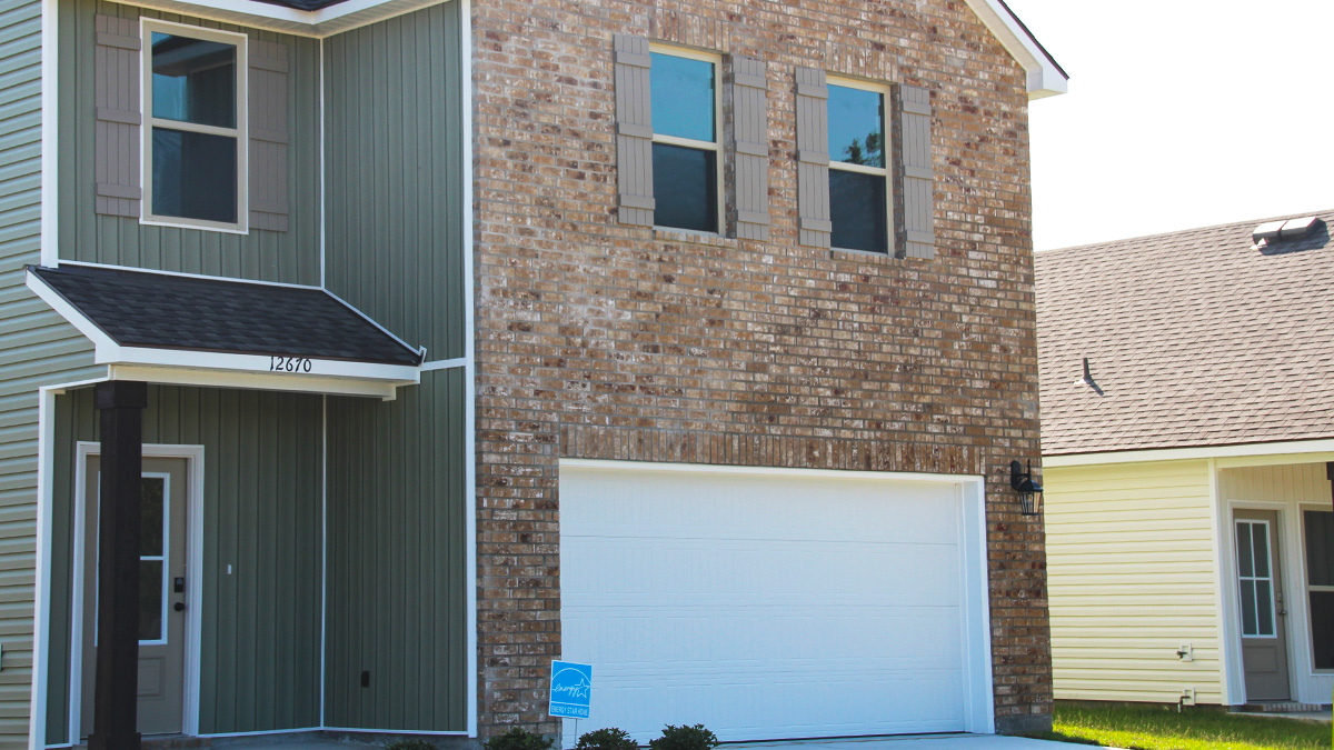 two story home with tan brick. Tan glass door and tan shutters. Green vinyl and white two car garage and long driveway.