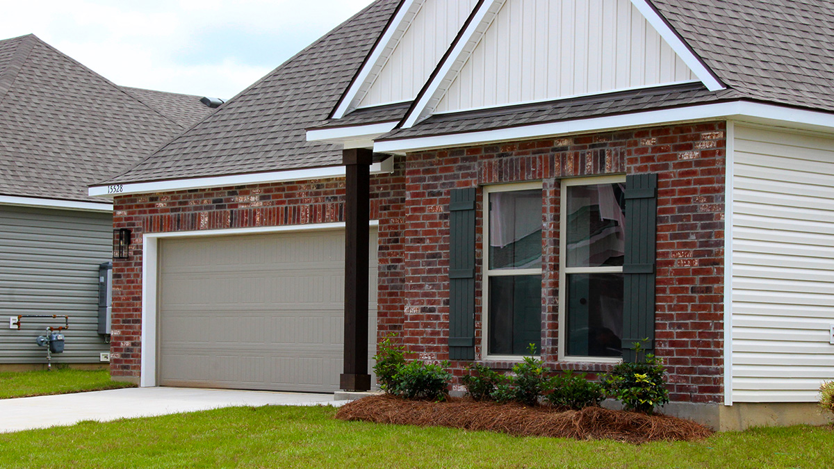 Single-story home with red brick. Black glass front door with dark grey shutters and tan two car garage.