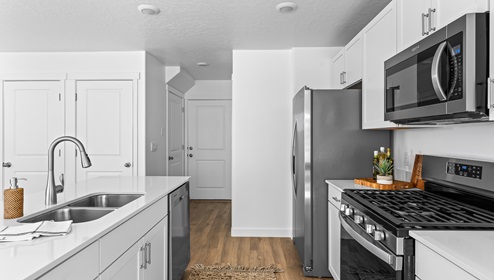 A wide entryway through the kitchen with white countertops and shaker cabinets, stainless steel appliances and a large island with a sink.