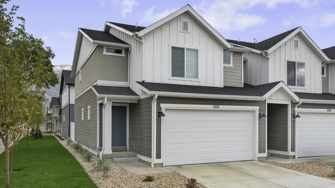 New two-story farmhouse style townhome in Toole Utah with taupe and white colored siding and white trim and a covered porch with a two car garage.