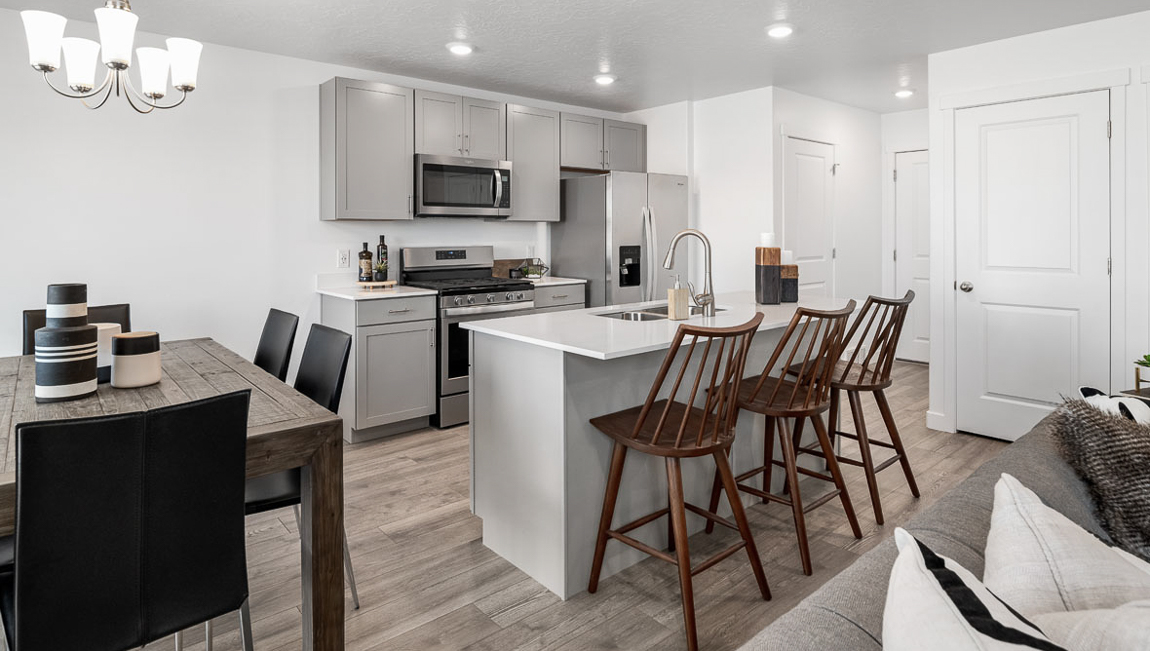 View of the living room leading into the dining area with a 6 seat table and kitchen with grey shaker cabinets, stainless steel appliances and breakfast bar with three stools.