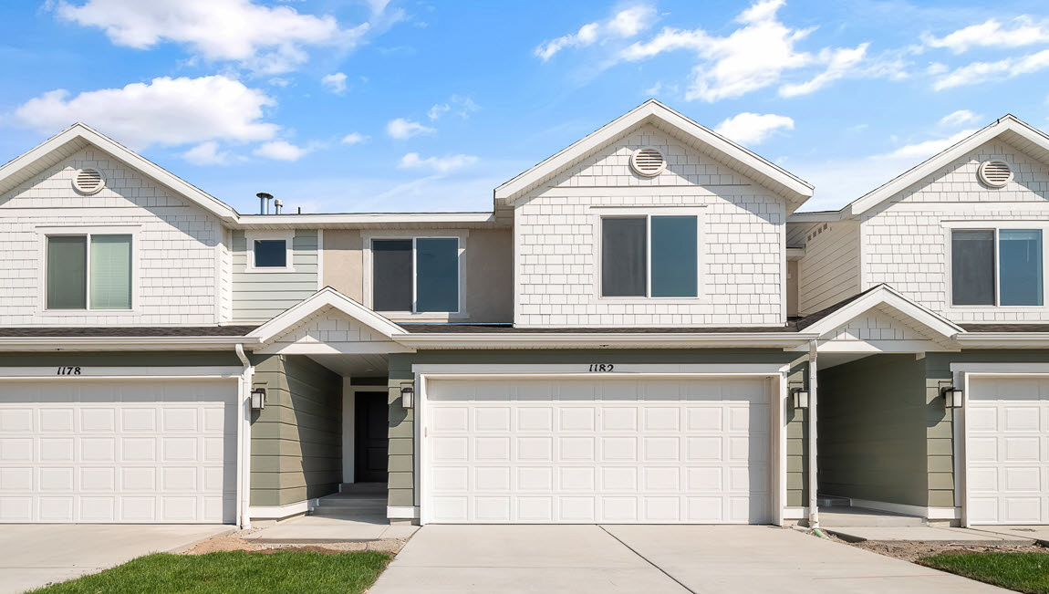 New two-story townhome in Tooele Utah with light sage green, light peach and cream colored siding and white trim and a covered porch with a two car garage.