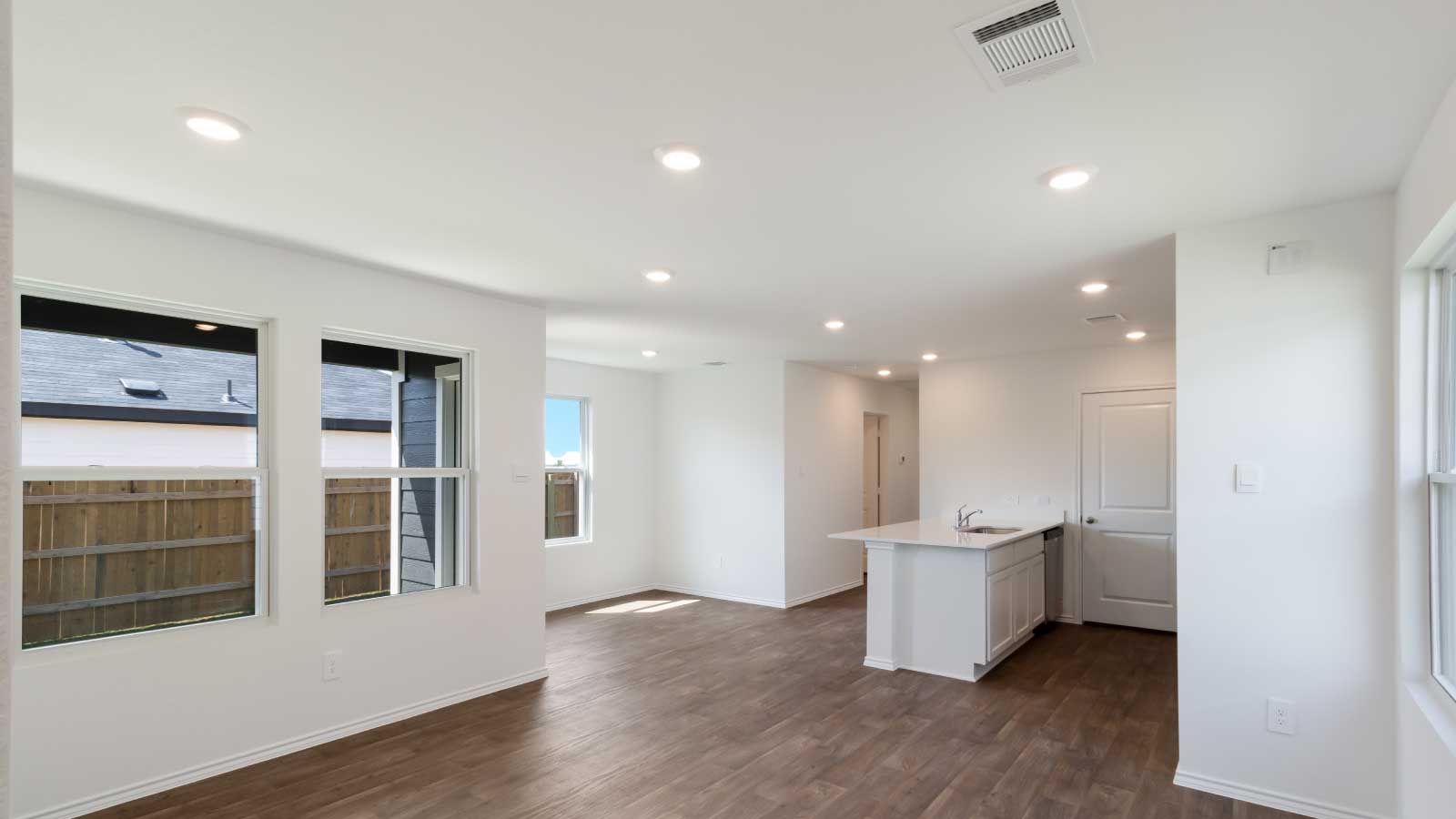Living room area connecting to kitchen with dark brown flooring and white walls.