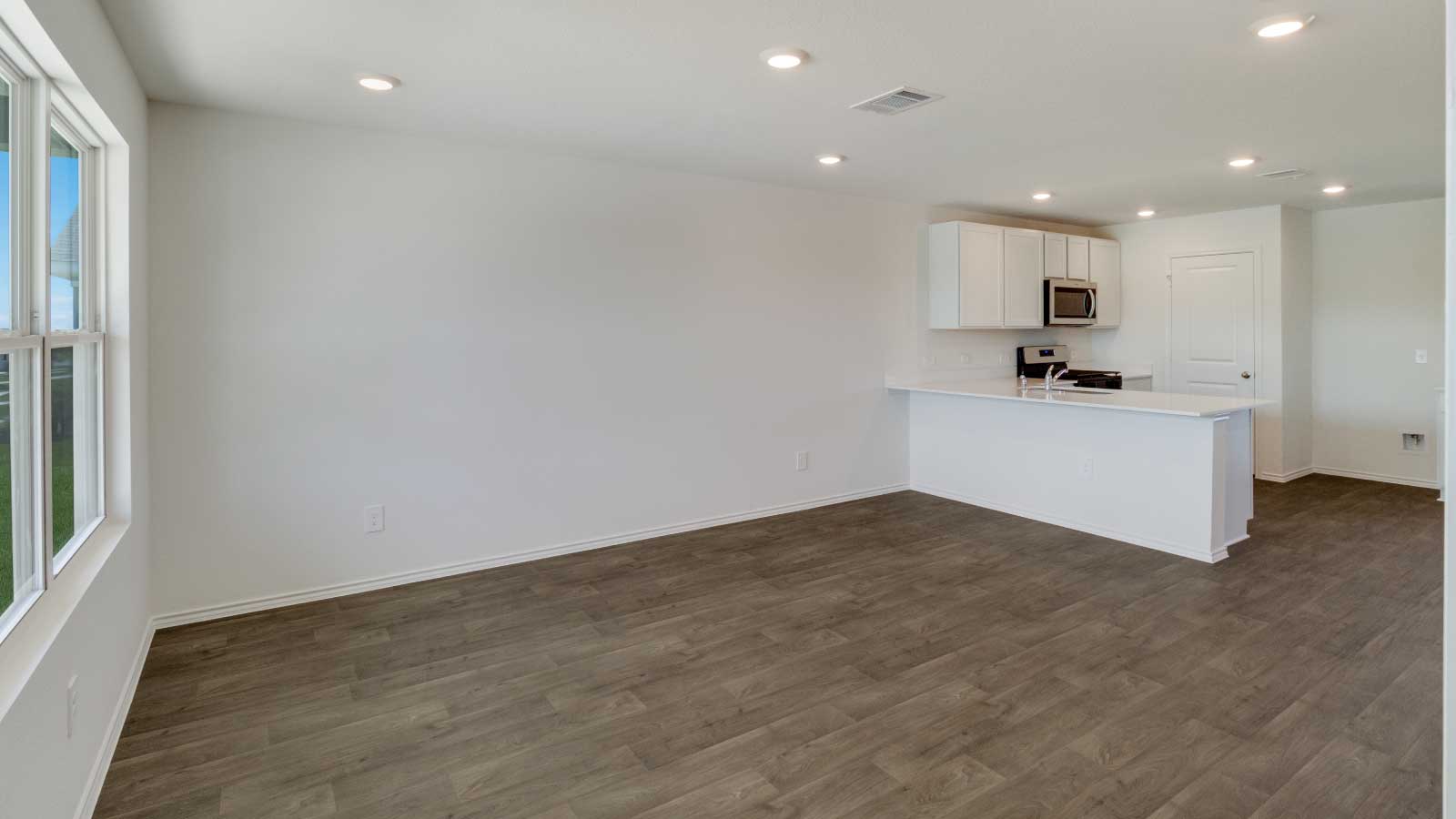 Living area with brown flooring and white walls connecting to the kitchen.