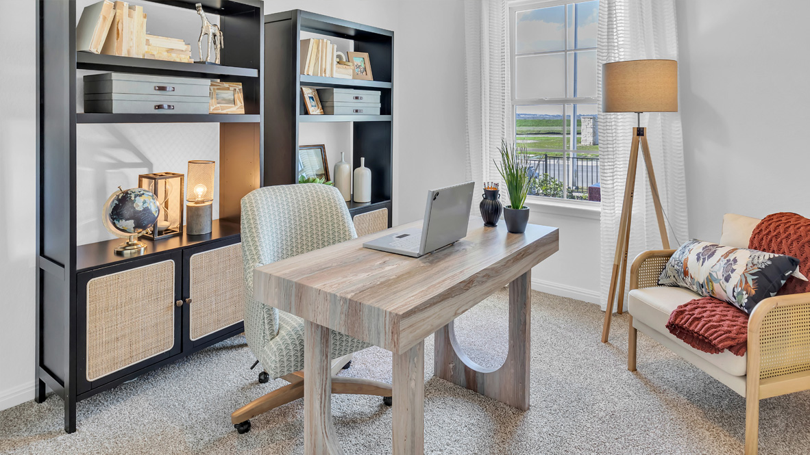 Cozy home office with a wooden desk, laptop, and a potted plant. Bookshelves hold decor and books. A lamp and armchair add warmth by a window.
