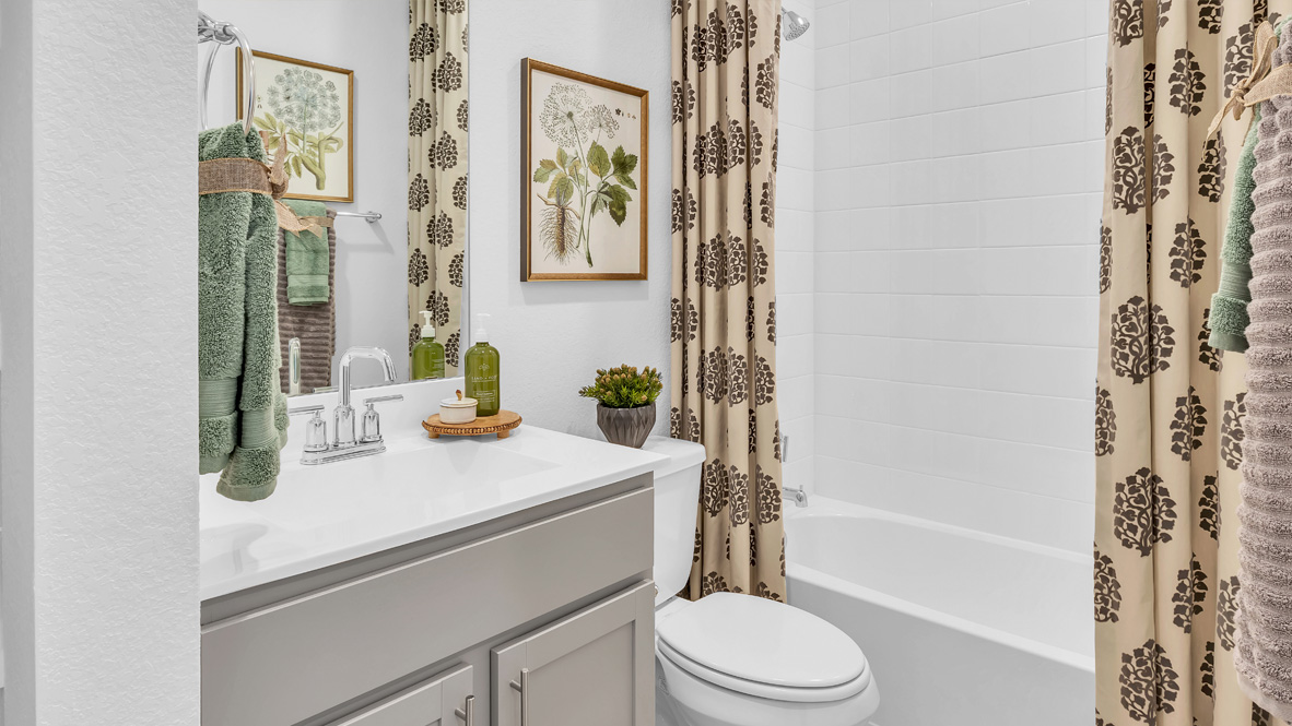 A bright bathroom with beige cabinets and green towels. Botanical prints and beige curtains with brown patterns add a natural, calming ambiance.