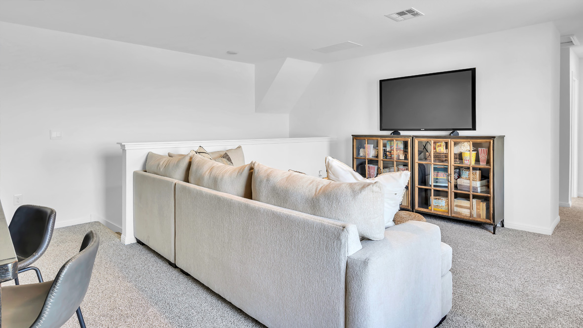 Comfortable living room with a light beige L-shaped sofa facing a large flat-screen TV on a cabinet filled with books and decor, set on a plush carpet.
