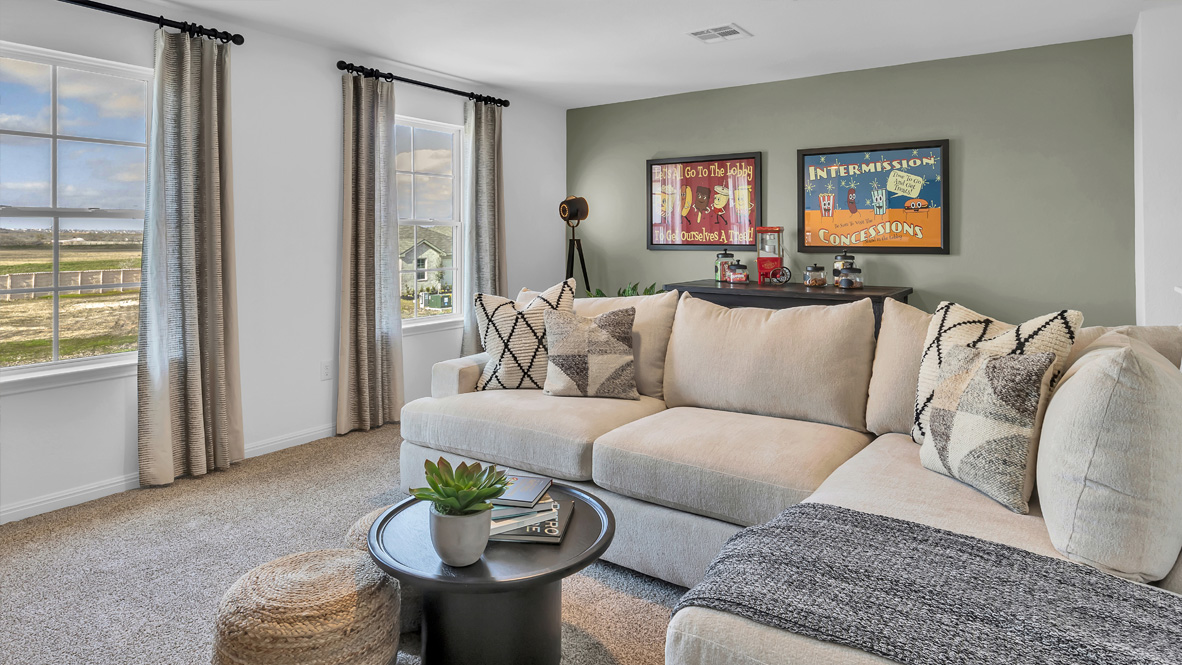Cozy living room with beige sectional sofa adorned with patterned pillows. Two framed posters, a plant, ottoman, and windows with scenic view enhance the space.