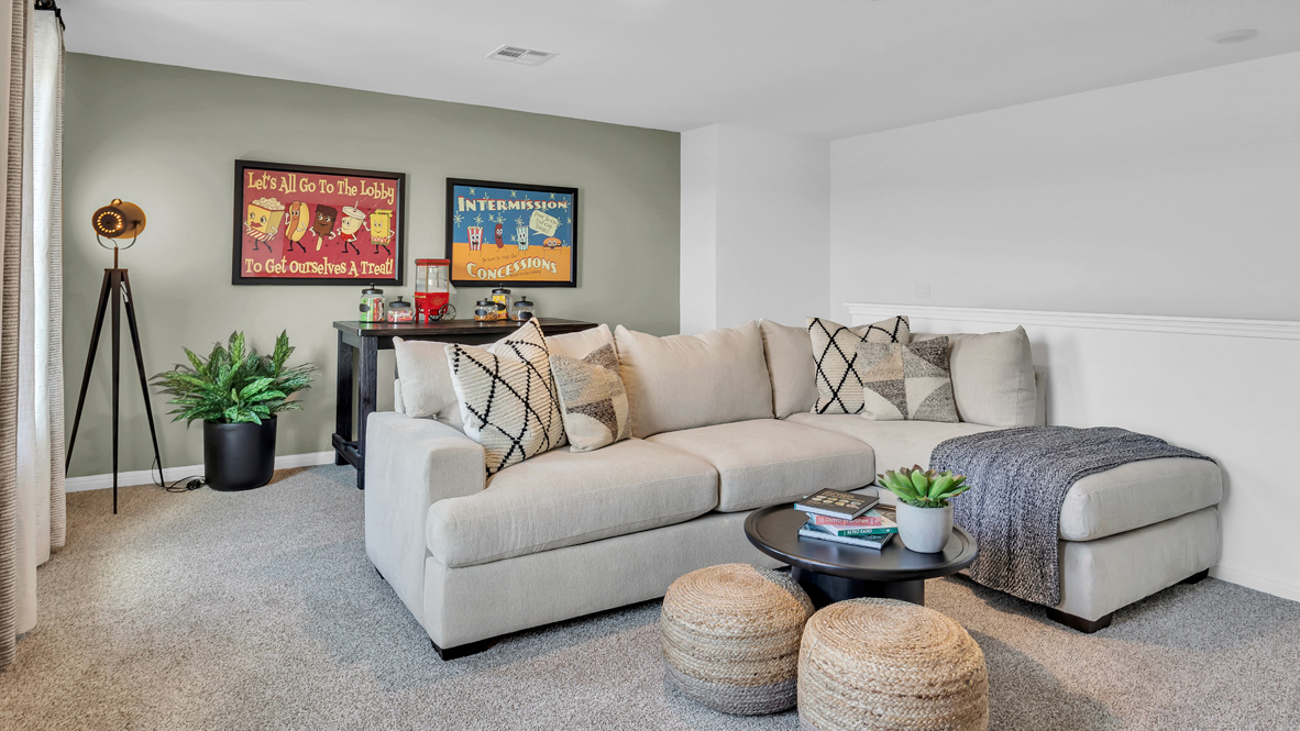 Cozy living room with a beige sectional sofa and geometric pillows. A gray throw blanket drapes over the couch. Two framed vintage posters hang above a black bar cart. A round black coffee table holds books and a plant. Two woven poufs add texture below. A tripod lamp and potted plant decorate the corner. Soft, inviting ambiance.