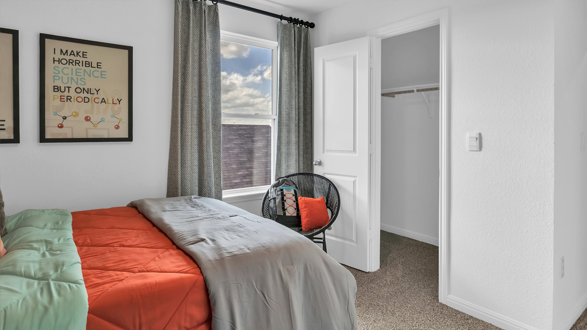 Cozy bedroom with an orange and gray bed, science pun wall art, and an open closet door. Black chair with colorful cushions near the window with gray curtains.