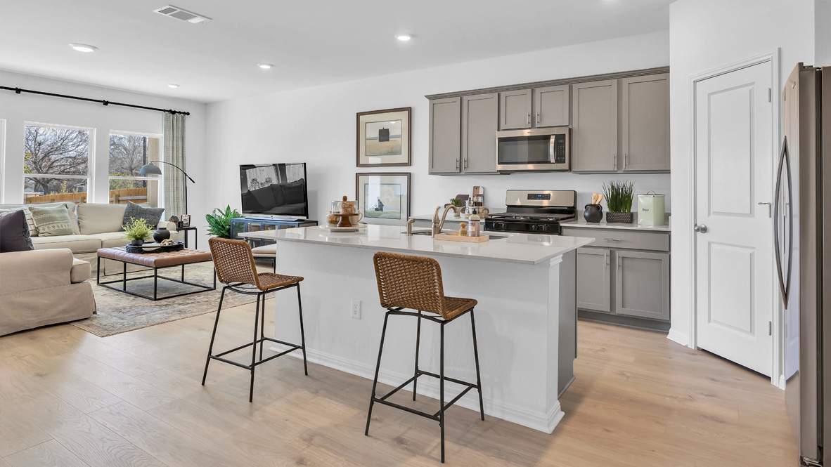 Modern kitchen and living room with light wood floors, gray cabinets, and stainless steel appliances. A cozy beige sofa faces a flat-screen TV, and two wicker stools stand by the kitchen island, creating a warm, inviting atmosphere.